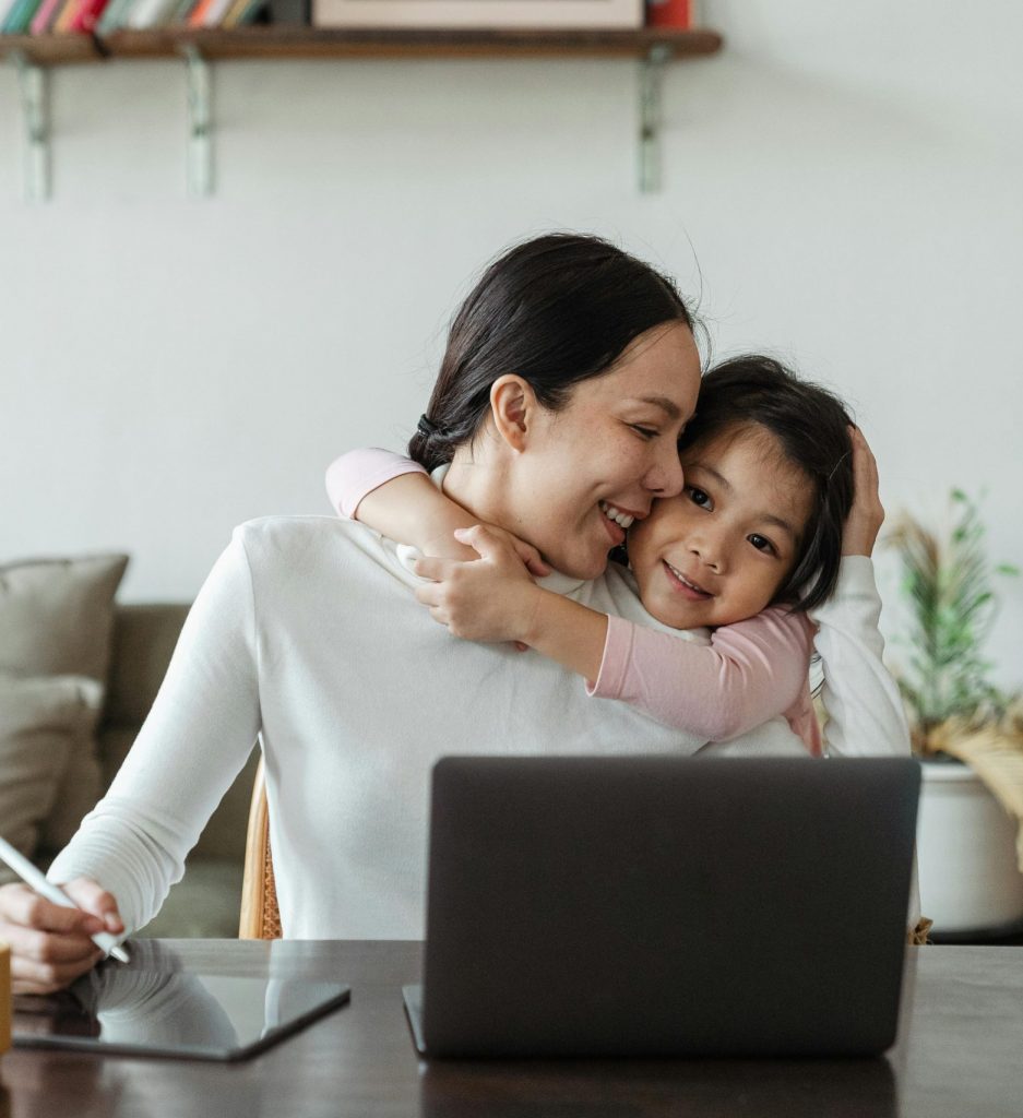 Happy young Asian woman working remotely from home with laptop and tablet while adorable little daughter hugging from behind